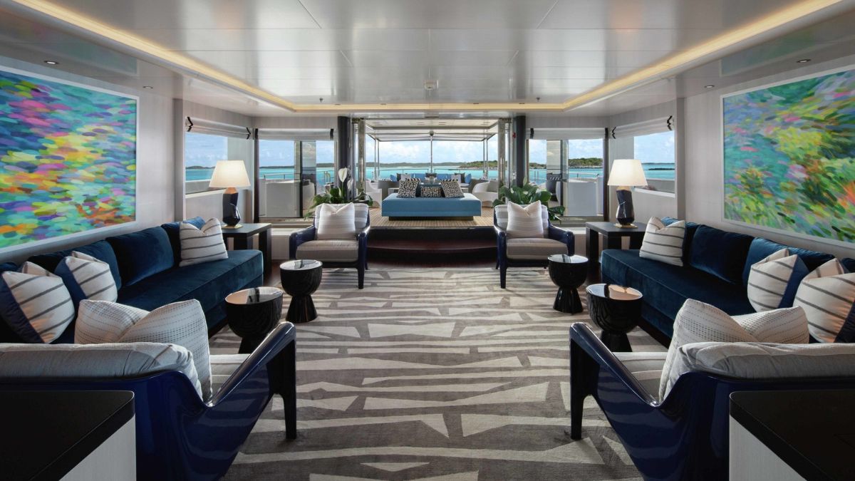 A stylish yacht interior featuring plush blue sofas, decorative pillows, and wooden side tables. Colorful abstract artwork adorns the walls, and large windows provide a view of the ocean. The space is bright and modern, with a neutral patterned rug on the floor.