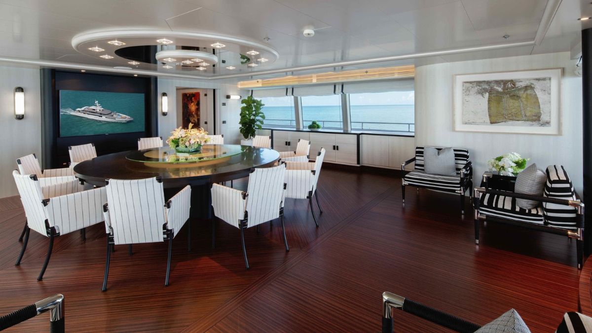 A spacious indoor dining area featuring a large round table surrounded by white chairs. Floor-to-ceiling windows offer a view of the ocean. Modern decor includes artwork and plants, with striped armchairs in the corner.
