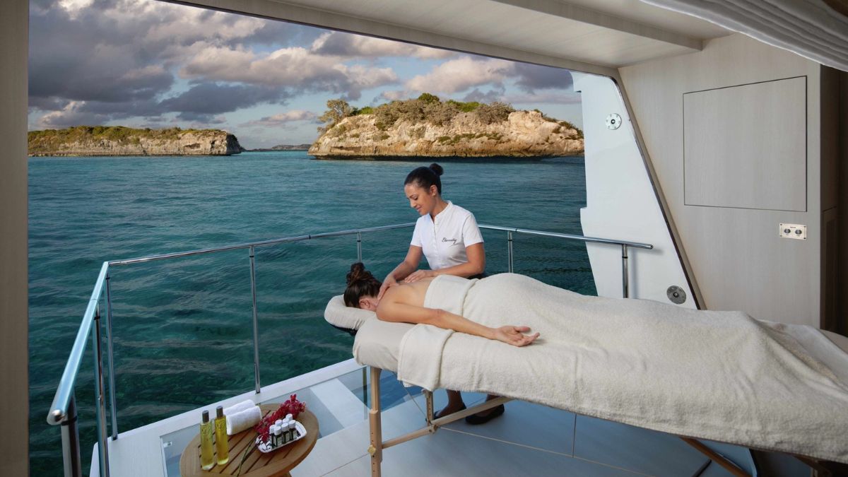A person receiving a massage on a balcony overlooking a calm ocean. Scenic islands are visible in the background under a partly cloudy sky. Spa products are neatly arranged on a small table nearby.