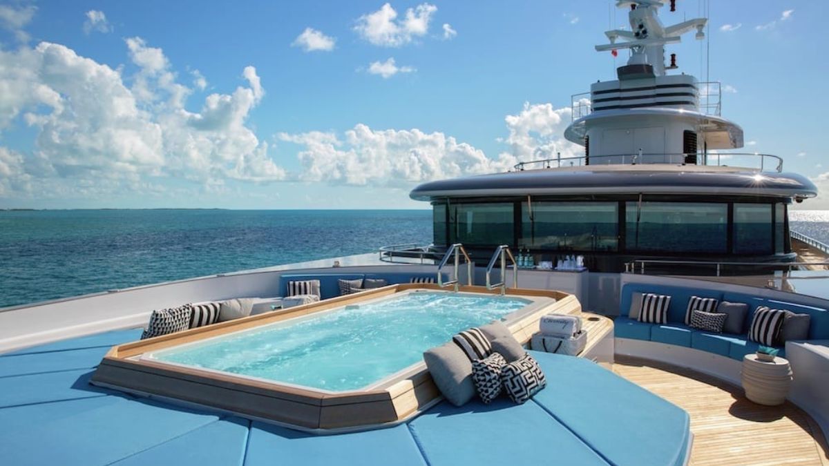 Luxury yacht deck featuring a whirlpool spa overlooking the ocean. Plush seating surrounds the pool, with bright blue skies and clouds in the background.