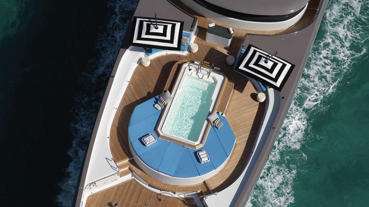 Luxury yacht deck featuring a jacuzzi surrounded by lounge chairs. Two striped umbrellas provide shade over the seating area. Clear turquoise water surrounds the vessel.