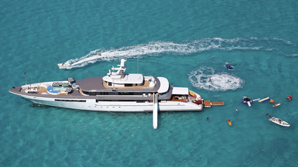 A luxurious yacht anchored in clear turquoise waters, surrounded by various watercraft, including jet skis and smaller boats. People are enjoying water activities nearby.