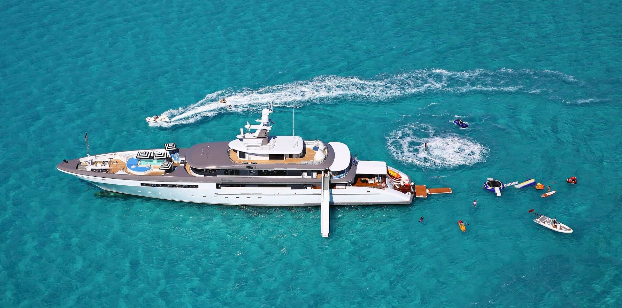 A luxurious yacht anchored in clear turquoise waters, surrounded by small boats and water sports activities. The yacht features multiple decks and a sleek design, while people engage in fun on the water nearby.