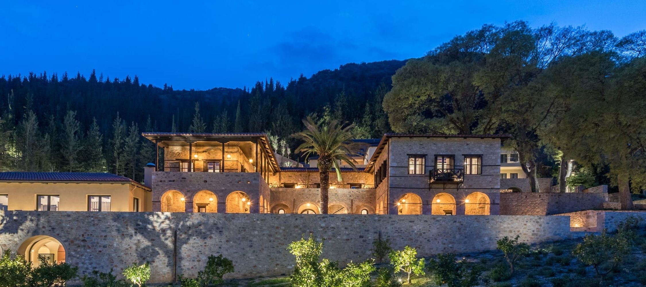 A beautifully lit stone villa at dusk, surrounded by lush greenery and trees. The architecture features arches and a mix of open and covered terraces, with warm lights glowing from the windows.