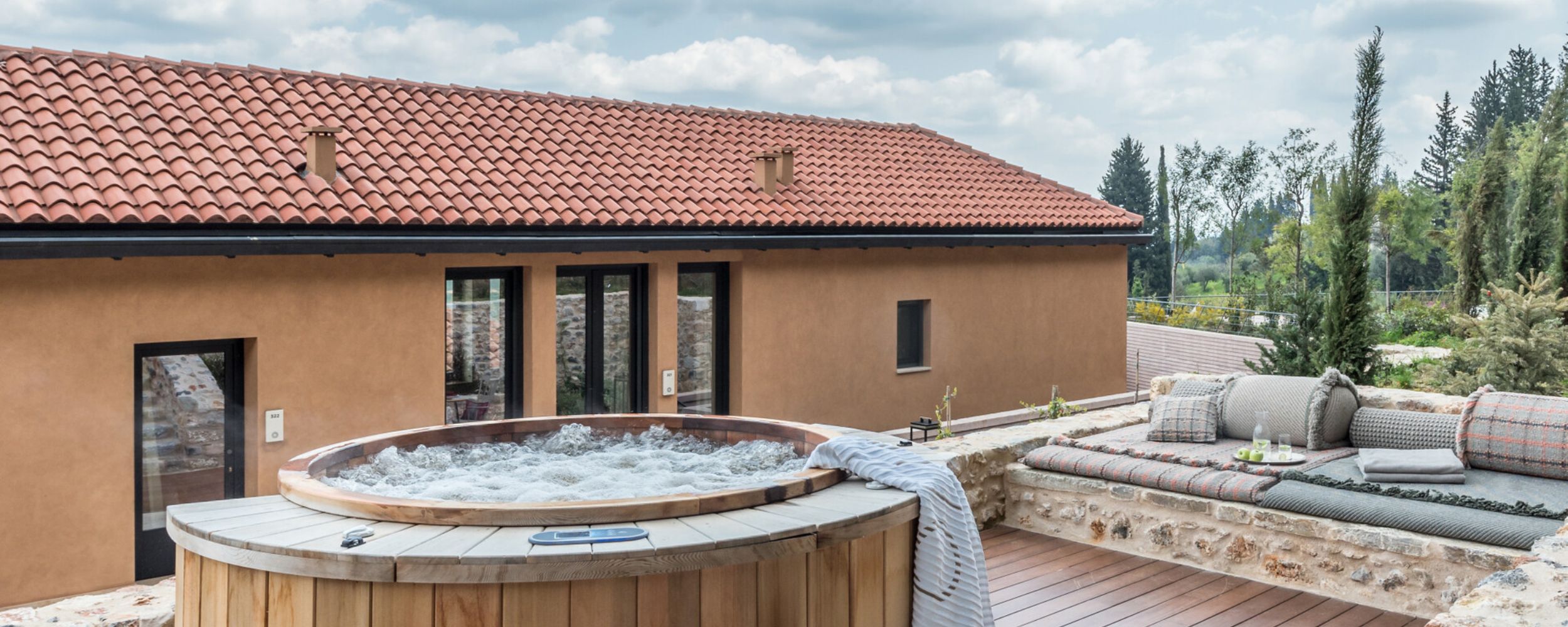 A wooden hot tub sits on a terrace with a stone wall, surrounded by comfortable outdoor seating on plush cushions. The backdrop features a building with a terracotta roof under a cloudy sky, with trees visible in the distance.