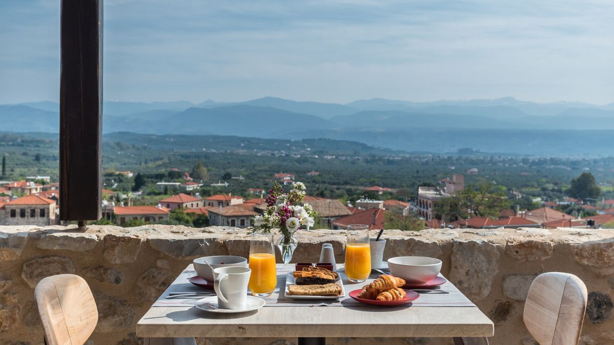 A beautifully set breakfast table with pastries, orange juice, and a small vase of flowers, overlooking a scenic view of rolling hills and distant mountains.