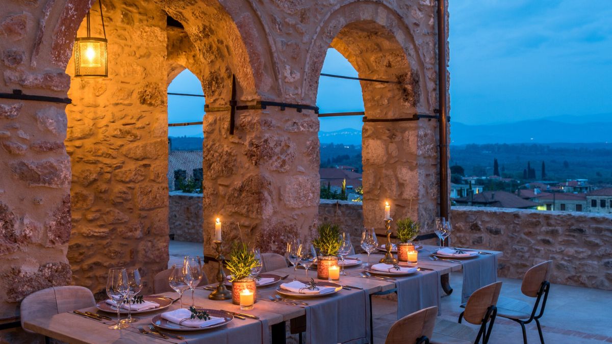 A dimly lit outdoor dining area featuring rustic stone arches, elegantly set tables with candles and greenery, overlooking a scenic landscape at dusk.