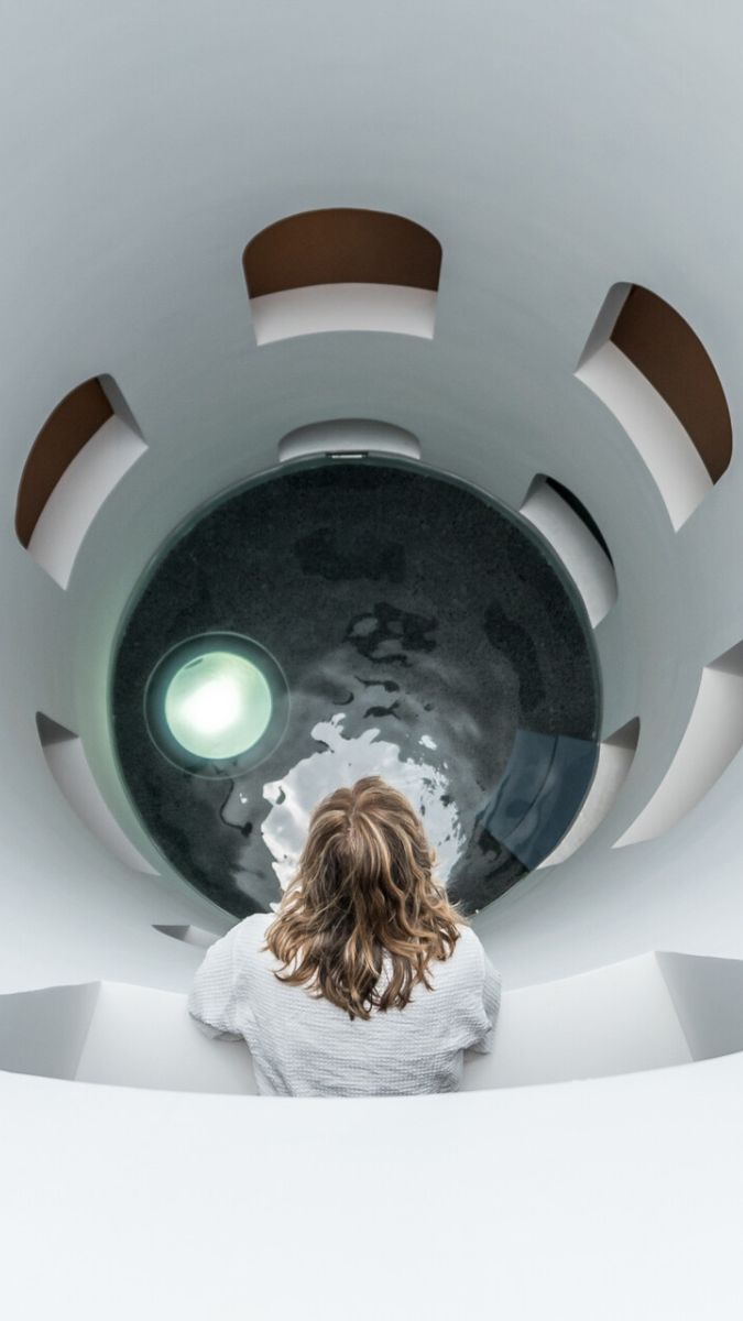 A person with long, wavy hair sits at the edge of a circular, well-lit opening that leads to a deep, reflective pool of water below. The smooth, white walls create a minimalist aesthetic, emphasizing the depth beneath.