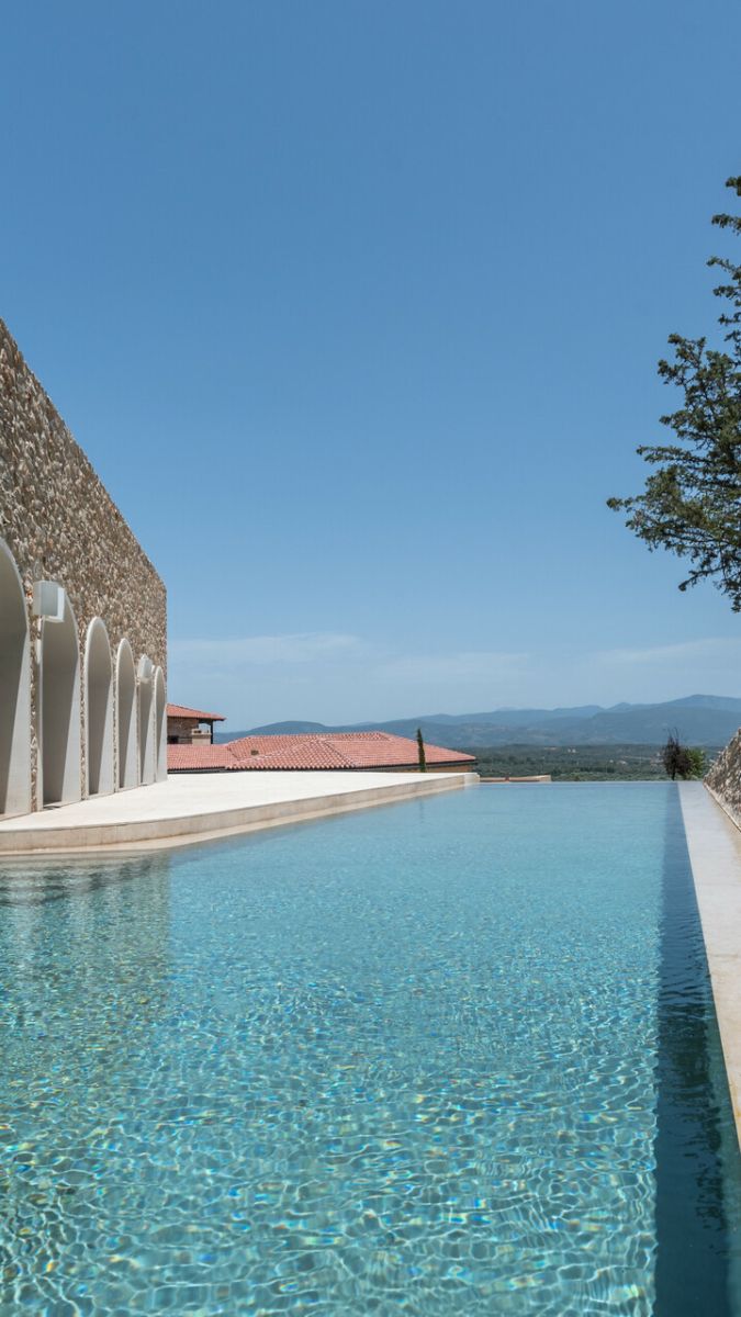 A tranquil infinity pool reflects the clear blue sky, surrounded by a stone wall and lush greenery. In the background, red-roofed buildings and distant mountains create a picturesque landscape.