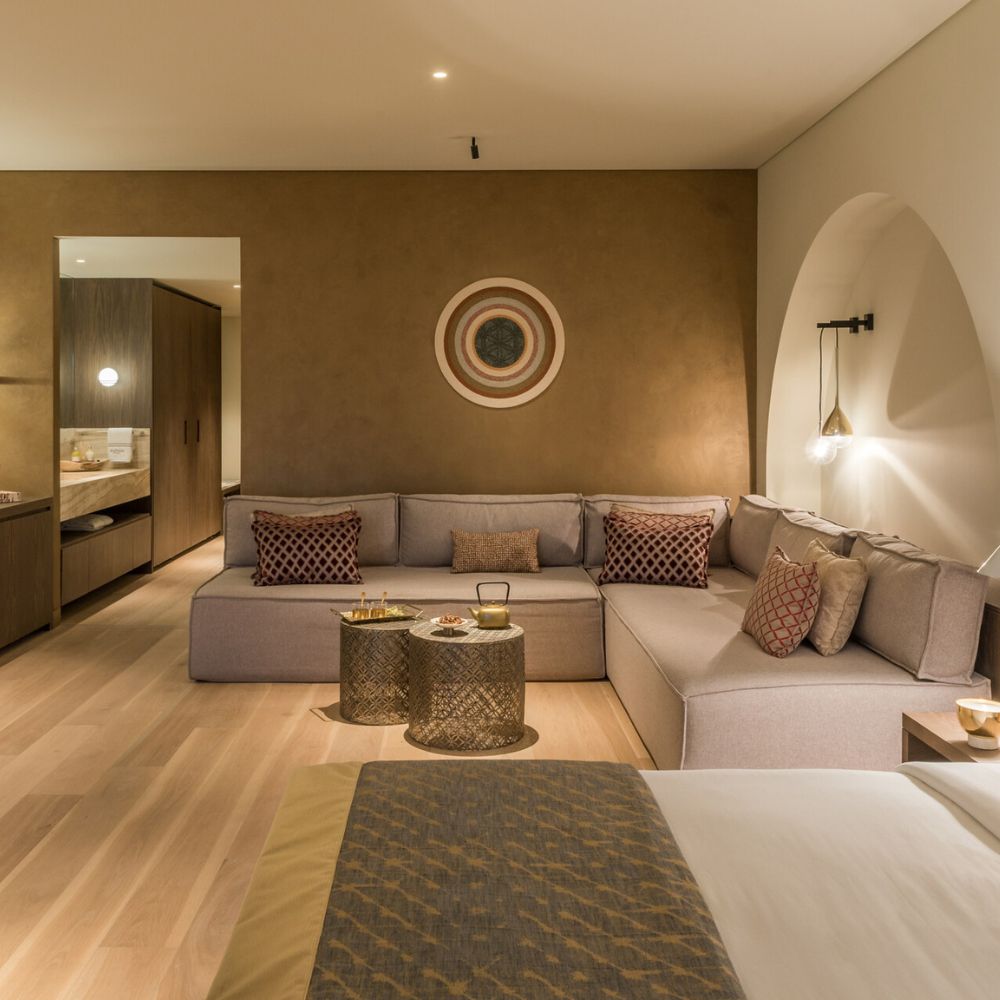 A stylish hotel room featuring a cozy seating area with a modern gray couch, decorative pillows, and two gold accent tables. The ambient lighting creates a warm atmosphere, complemented by earthy brown walls and a minimalist decor.