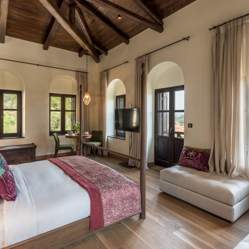 A spacious and elegantly decorated bedroom featuring a cozy king-sized bed with a richly colored throw, wooden beams on the ceiling, and large windows that allow natural light to fill the room. A small sitting area with a sofa is present, along with a desk and chair by the window.