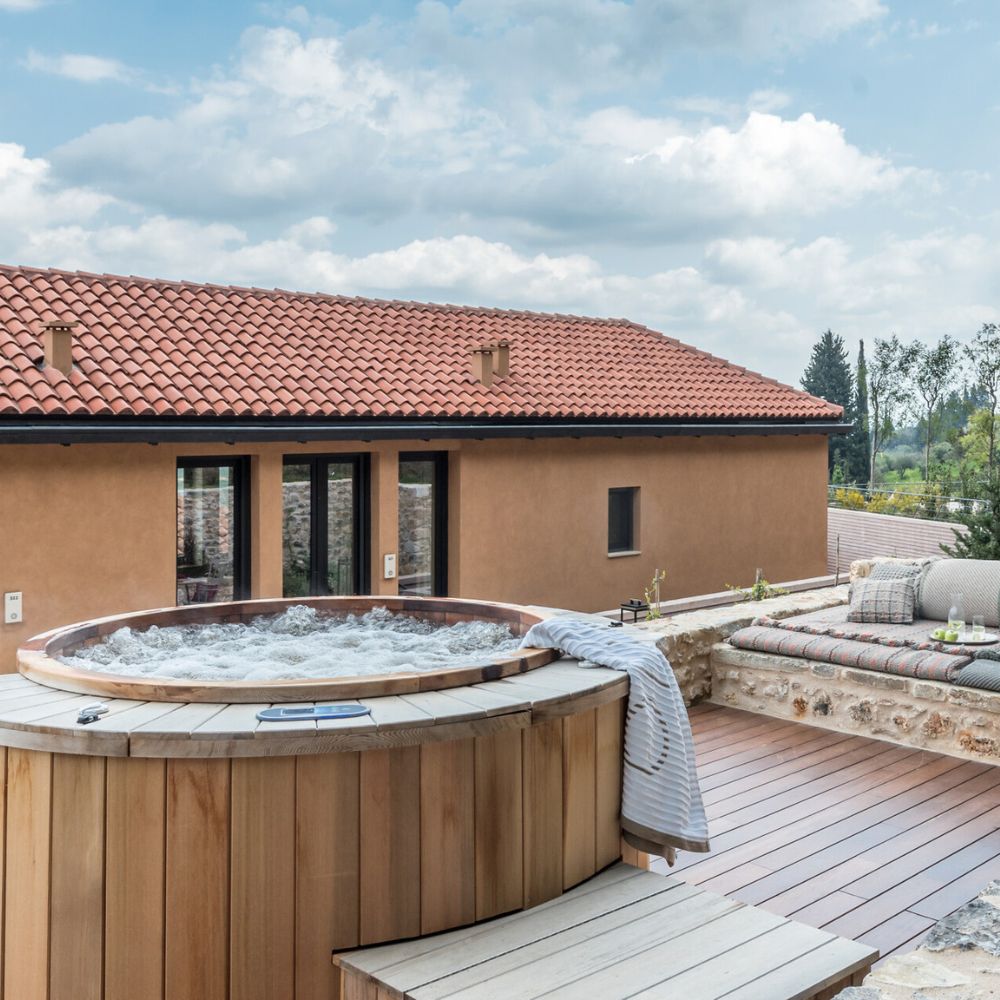 A wooden hot tub sits on a deck, surrounded by a stone wall, with a cozy seating area featuring cushions nearby. In the background, a house with a terracotta roof and large glass doors is visible under a partly cloudy sky.