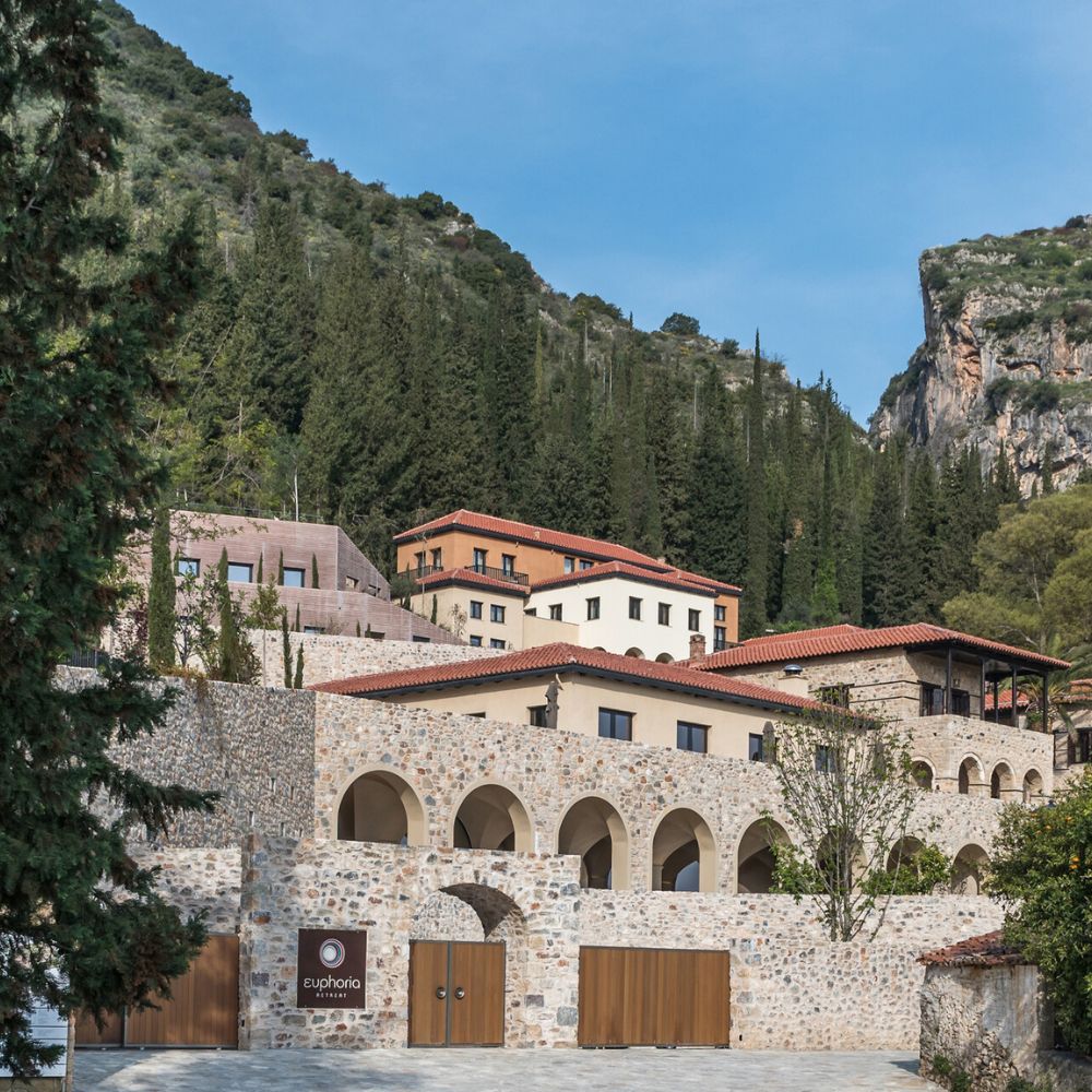 A charming stone building with arched entrances, surrounded by lush greenery and mountains. The structure features a mix of modern and traditional architecture, with red tile roofs and a welcoming sign that reads "Euphoria."