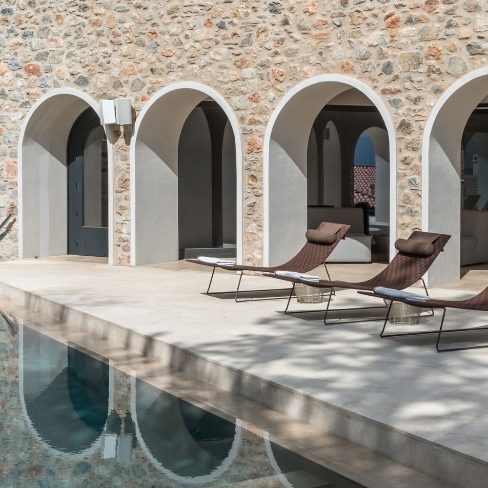 A modern, stone-walled villa with arched doorways opening to a lounge area. Two sleek sun loungers are positioned beside a crystal-clear swimming pool, reflecting the serene surroundings.