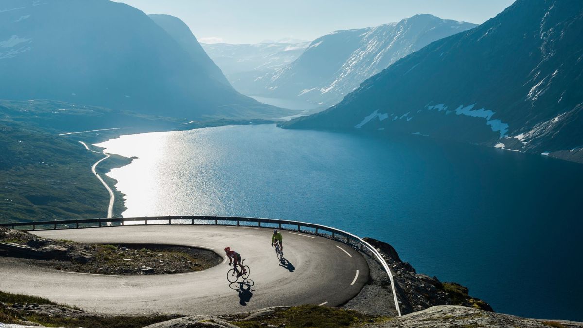 Two cyclists ride along a winding mountain road with a stunning view of a fjord and snow-capped mountains in the background. The sun shines brightly, reflecting off the water.