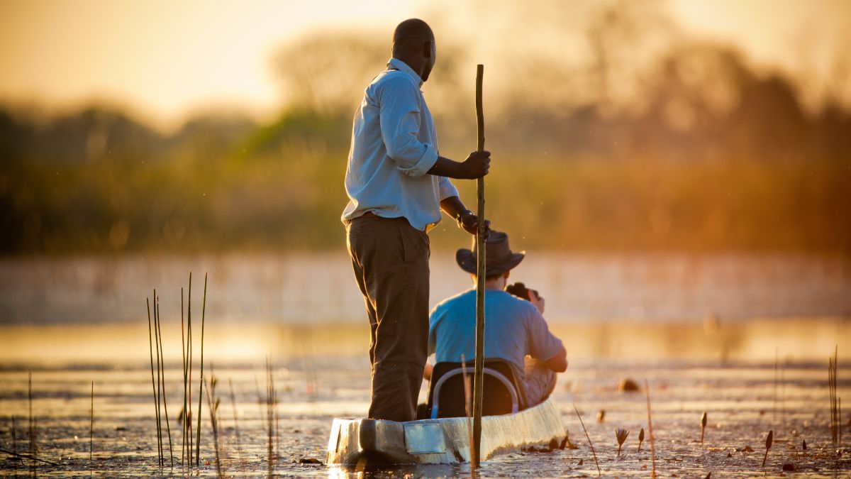 Two people in a canoe navigating through a tranquil waterway during sunset. One person is standing and maneuvering a long pole, while the other is seated and taking photographs. Lush greenery and soft light create a serene atmosphere.