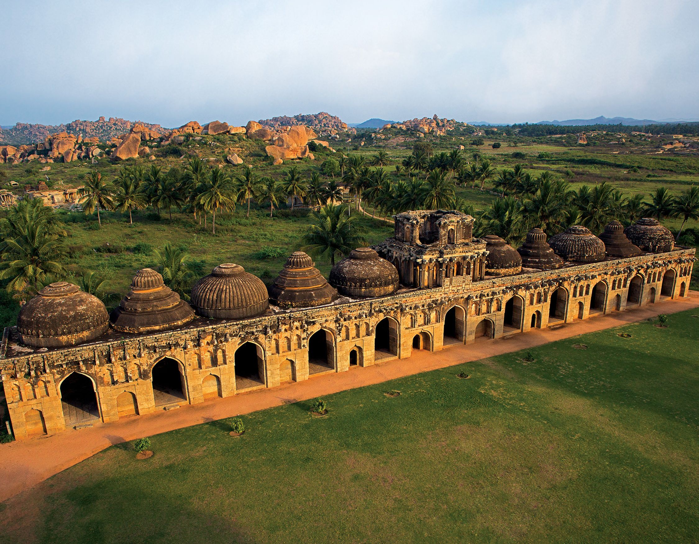 Aerial view of ancient stone architecture featuring multiple domed structures, set amidst lush greenery and rocky hills. Palm trees line the foreground, creating a serene landscape.