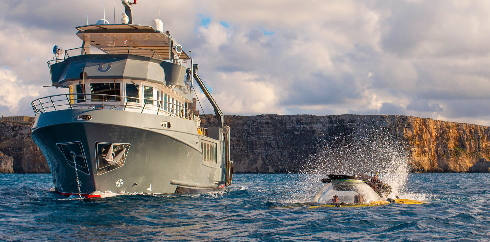 A large gray research vessel is anchored in the ocean, with a smaller yellow submersible nearby, creating splashes as it surfaces. Dramatic cliffs are visible in the background under a partly cloudy sky.