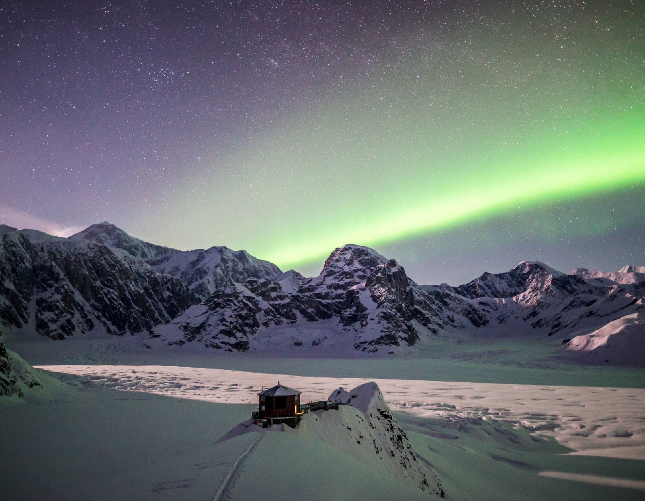 Green Northern Lights dancing over snowy mountains, viewed from a luxury private lodge in Iceland.
