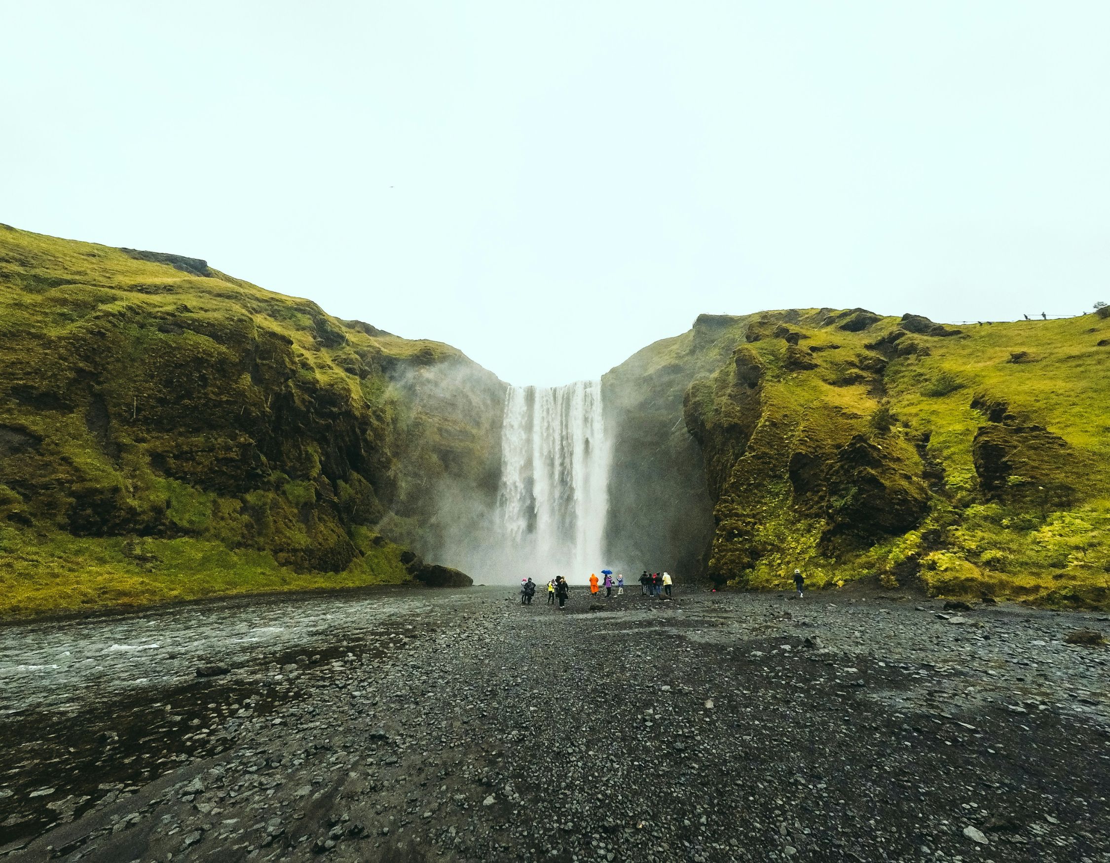 A stunning waterfall cascades down a rocky cliff surrounded by lush green hills. A group of people stands near the base of the waterfall, creating a sense of scale against the powerful flow of water. Mist rises from where the water meets the ground.