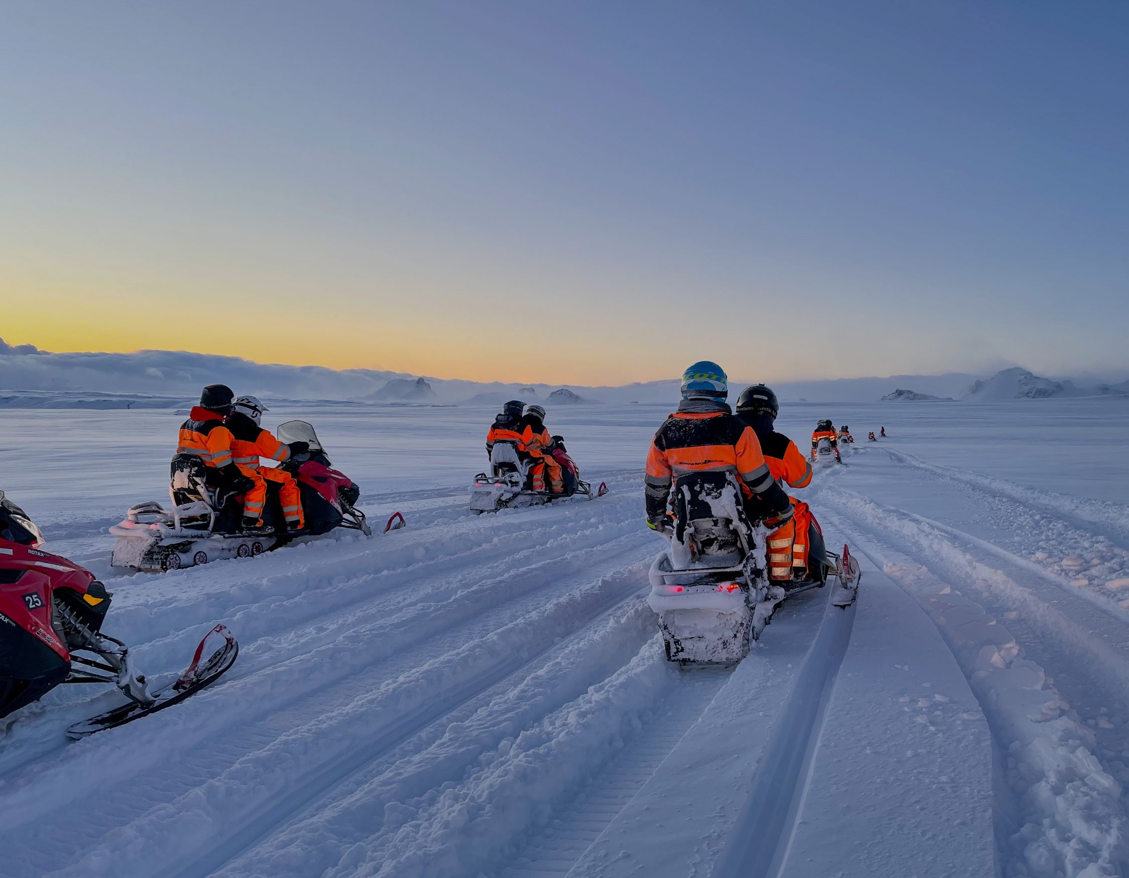 A group of snowmobiles rides across a vast snowy landscape during sunset. The riders wear bright orange winter gear, and the horizon is tinged with soft hues of orange and blue. Snow-covered mountains are visible in the distance.