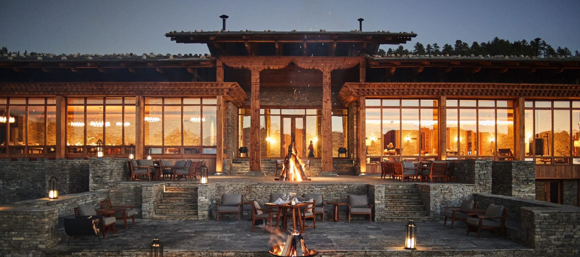 A beautifully designed restaurant with large glass windows, featuring a stone facade and wooden beams. The exterior has a terrace with tables and chairs arranged around a fire pit, creating a warm, inviting atmosphere at dusk.