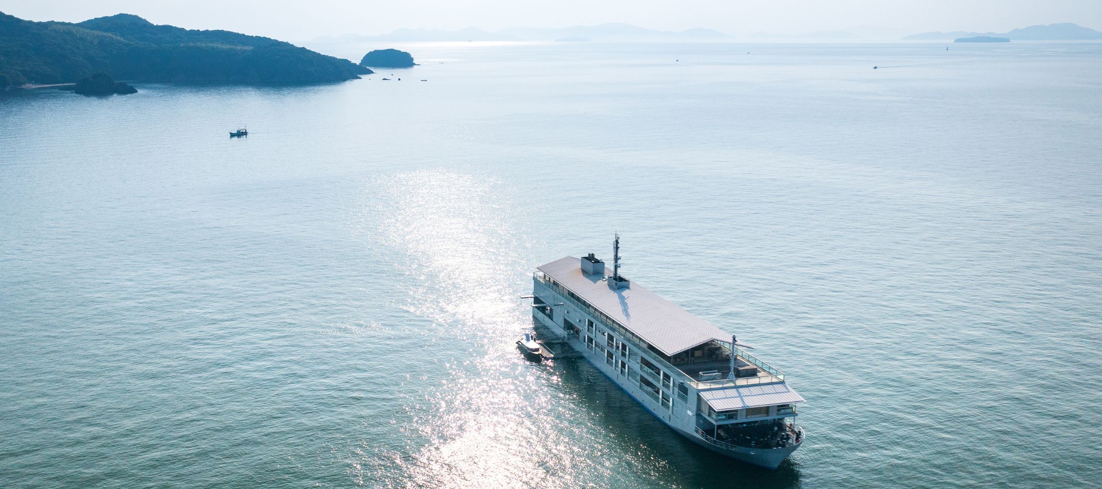A large ferryboat sails through calm, sunlit waters, with distant mountains and islands visible in the background. The surface of the water reflects shimmering sunlight.
