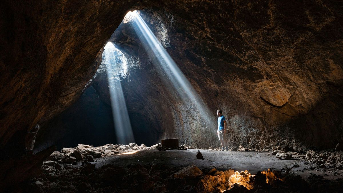 A person standing inside a cave, illuminated by beams of sunlight streaming through a hole in the cave ceiling. The rocky interior is dim, with textured walls and uneven ground.