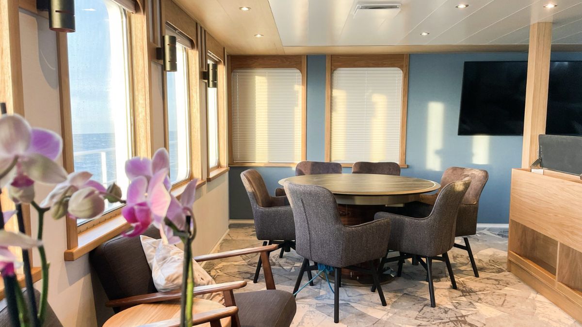 A bright, modern dining area with large windows overlooking the ocean. A circular wooden table is surrounded by comfortable, upholstered chairs. A few decorative potted orchids add a touch of elegance to the space.