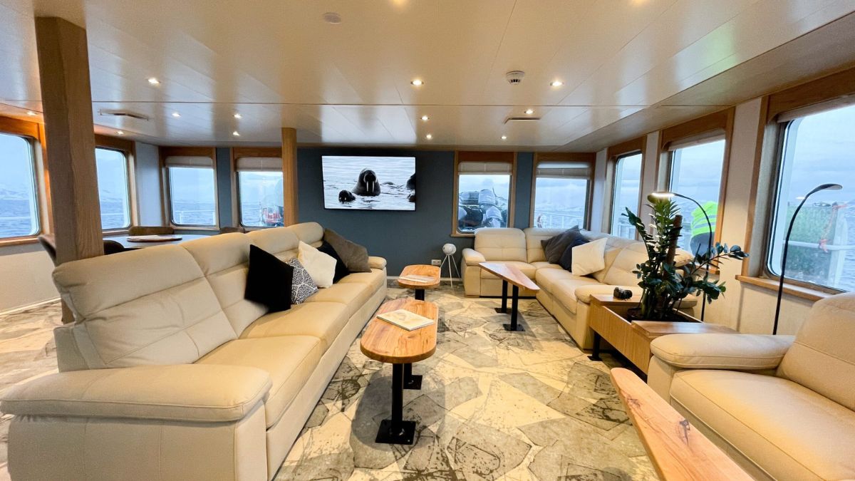 A cozy lounge area with cream leather sofas and wooden side tables, featuring a large screen displaying nature scenery. Large windows provide a view of the outside, and decorative plants add a touch of greenery to the space.