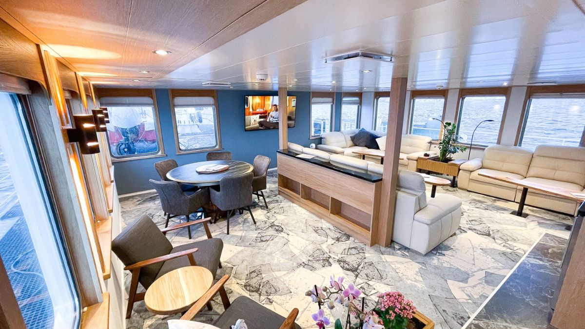A spacious interior of a modern boat, featuring a comfortable seating area with large windows overlooking water. The design includes a round dining table, stylish chairs, and warm lighting, creating a cozy atmosphere.