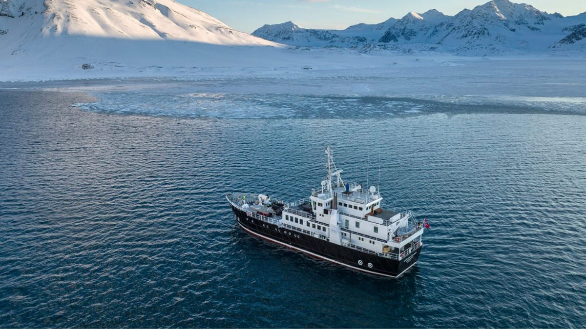 A research vessel navigates through icy waters surrounded by snow-covered mountains. Ice floes are scattered in the foreground, while the sun casts a soft light over the scene.