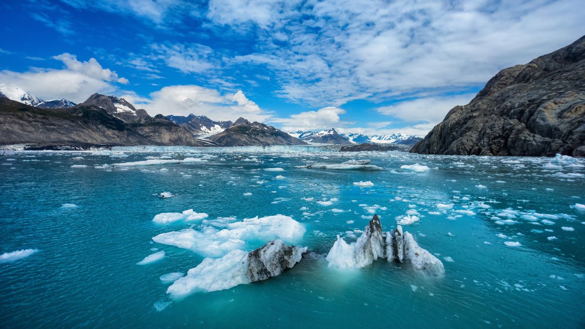 A serene glacial scene featuring floating icebergs in turquoise waters, surrounded by rugged mountains under a partly cloudy blue sky.