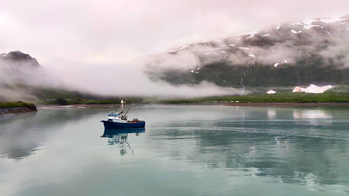 A blue fishing boat floats on calm water, surrounded by mist and mountains in the background. Snow-capped peaks are partially obscured by clouds, creating a serene and tranquil scene.
