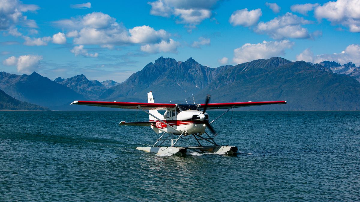 A small seaplane with red and white stripes is floating on calm water, surrounded by mountainous terrain and a bright blue sky with fluffy clouds.