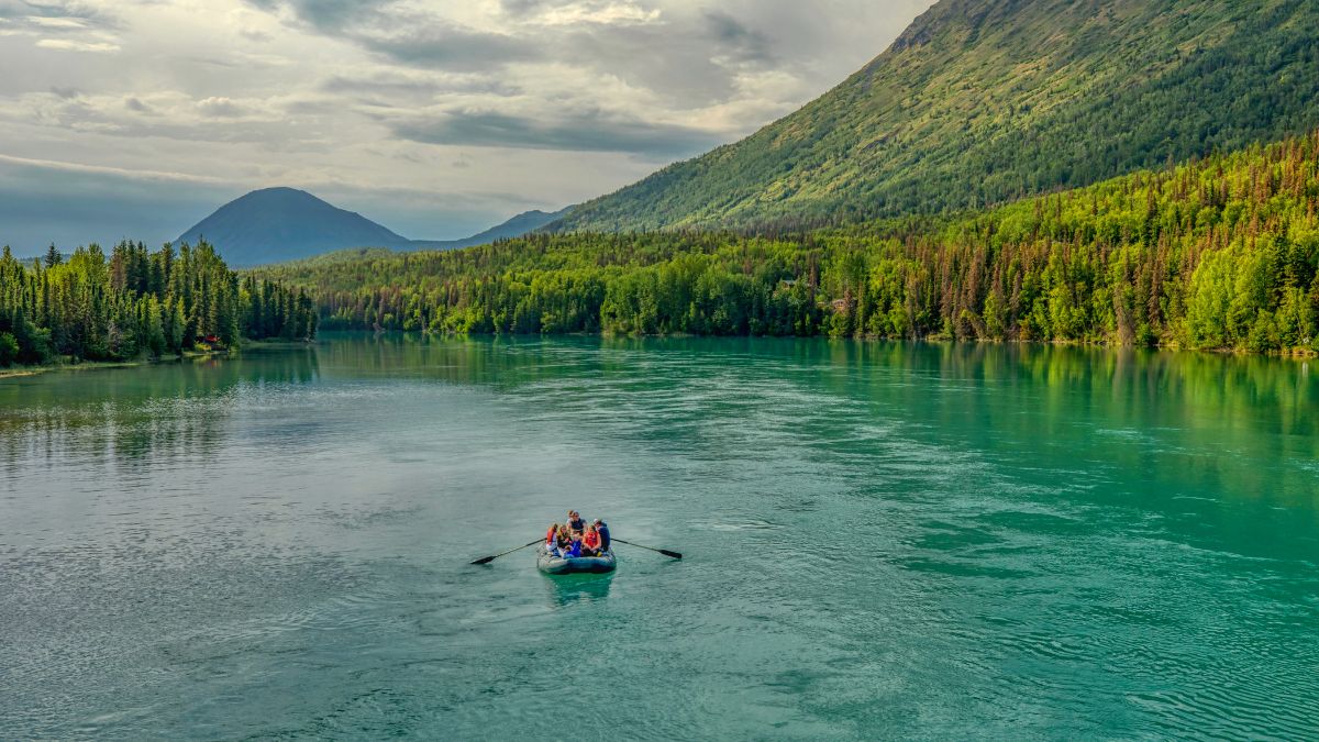 A group of people paddles in a small raft on a tranquil, turquoise river surrounded by lush green forests and mountains under a cloudy sky.
