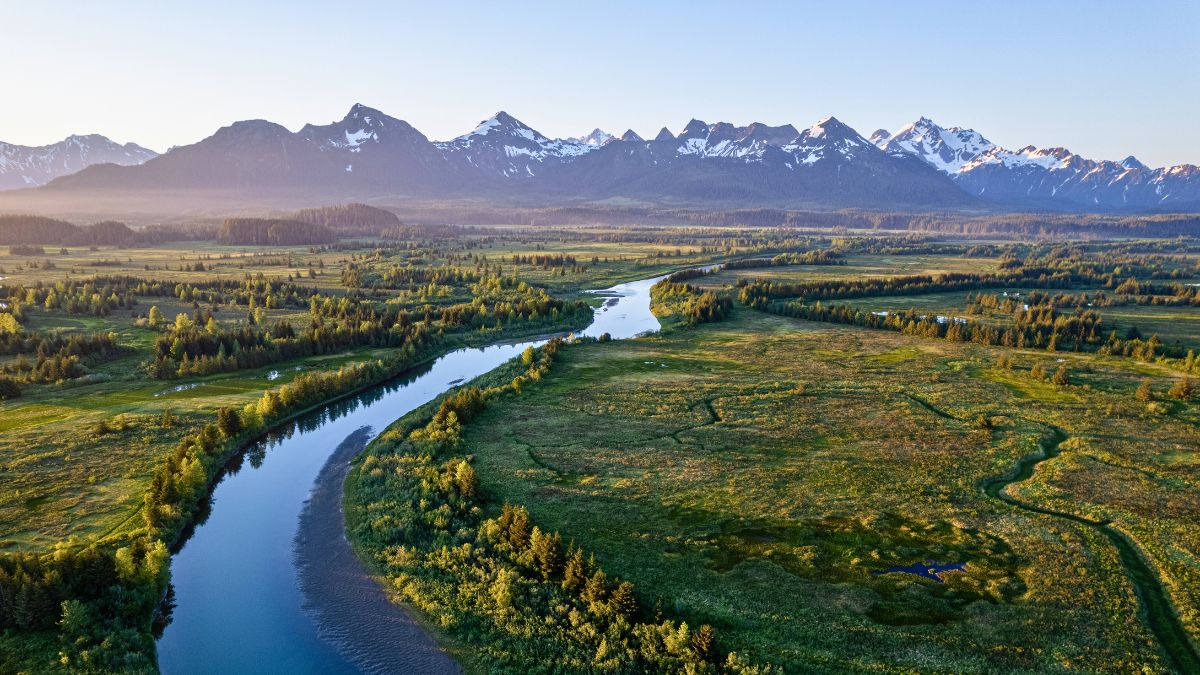A winding river flows through a lush valley surrounded by dense forests, with snow-capped mountains rising majestically in the background under a clear blue sky.