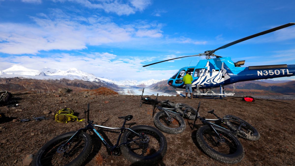 A blue and white helicopter is parked on rocky terrain with bicycles on the ground nearby. Snow-capped mountains and a glacier are visible in the background under a bright blue sky. A person is seen preparing gear next to the helicopter.