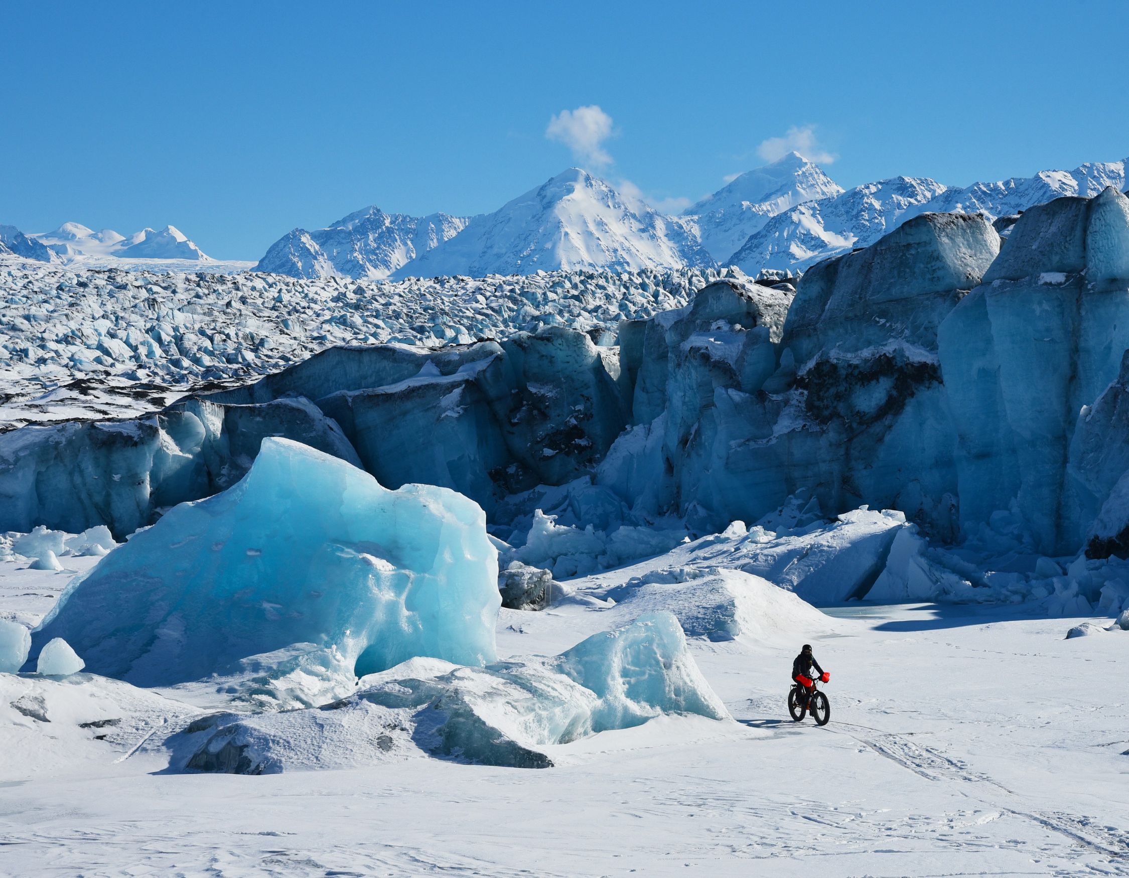 A person rides a fat bike across a frozen landscape, surrounded by large icy formations and snow-covered mountains under a clear blue sky.