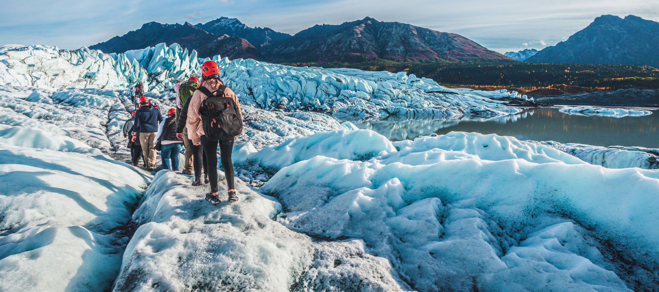 A group of people wearing helmets navigates a glacial landscape, with bright blue ice formations and rugged mountains in the background. The scene captures the beauty of nature in a wilderness setting.