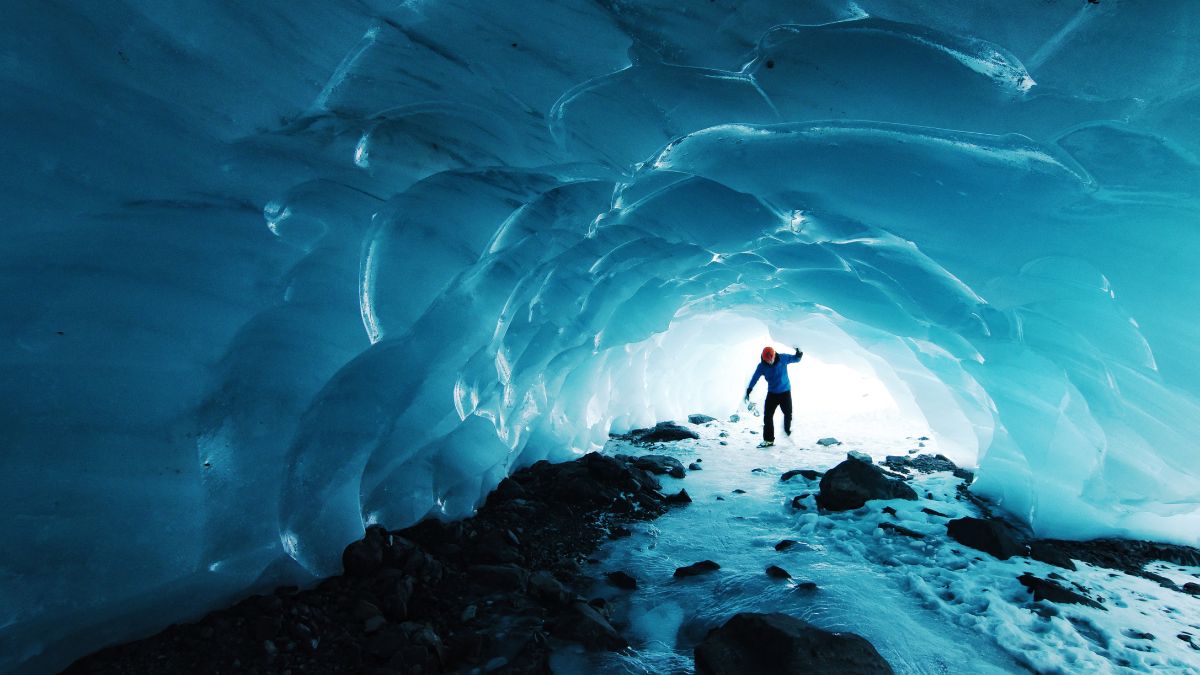 A person stands inside a blue ice cave, illuminated by light at the exit. The icy walls arch overhead, creating a stunning natural tunnel, with rocky ground visible beneath.