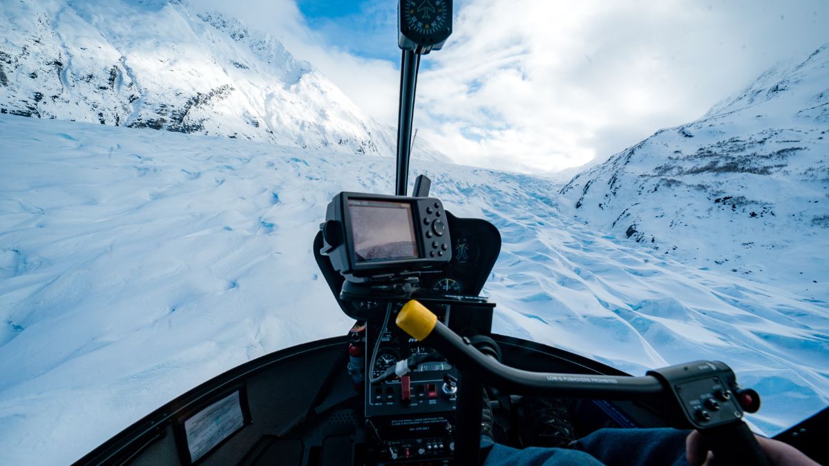 View from the cockpit of a helicopter flying over a snowy glacier landscape, showcasing vast ice formations and towering mountains under a partly cloudy sky.