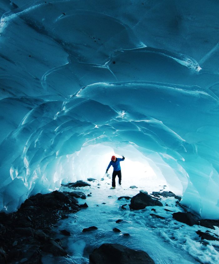 A person stands inside a glacial cave, illuminated by a bright light at the entrance. The icy walls glow in shades of blue, and water flows over rocky ground. The figure, wearing a bright orange helmet, reaches out toward the light.
