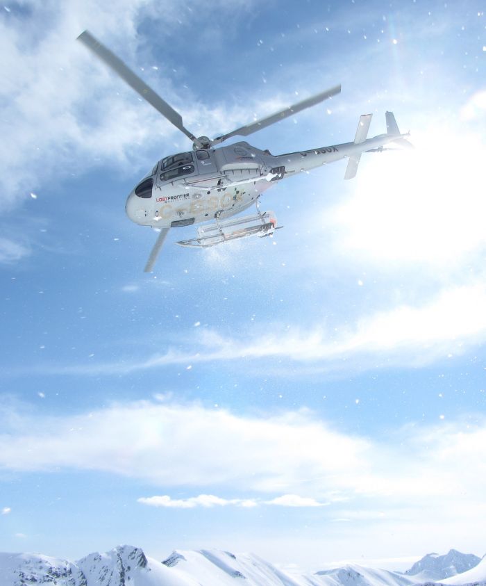 A helicopter flying over snow-covered mountains under a bright blue sky with scattered clouds. Sunlight reflects off the helicopter, creating a bright halo effect. Snowflakes are visible in the air.