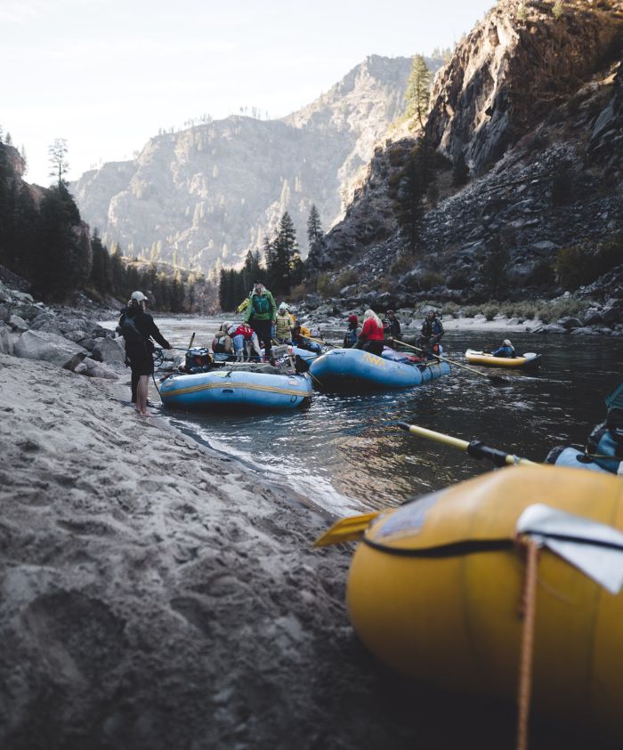 A group of people preparing rafts on a sandy riverbank surrounded by rocky mountains and pine trees. Some individuals are loading gear onto blue rafts, while others are standing or sitting around. The scene captures a sunny day with clear skies.