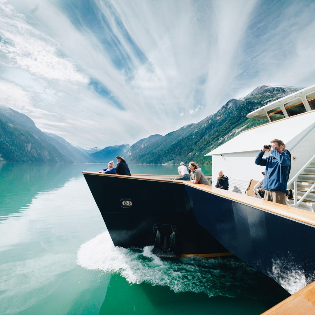 A group of people enjoying a boat ride on a calm, turquoise lake surrounded by mountains and dramatic clouds in the sky. Some are taking photos, while others admire the scenery.