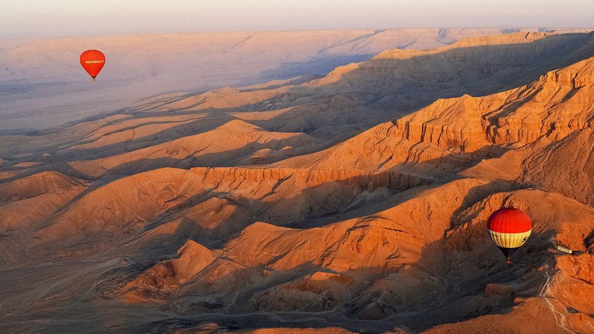 Hot air balloons float over rugged, orange-hued desert mountains at sunrise, casting gentle shadows on the landscape below.