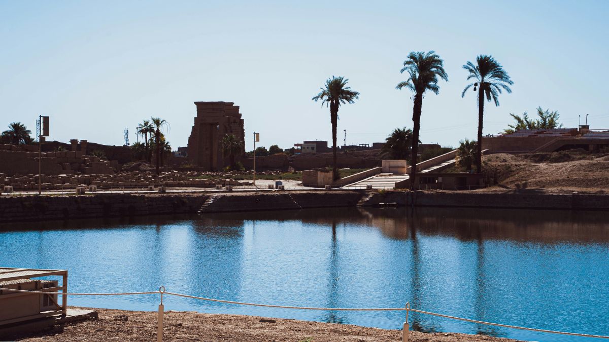 A tranquil scene featuring a body of water reflecting palm trees and ancient structures in the background. The sky is clear and blue, highlighting the serene atmosphere of the location.