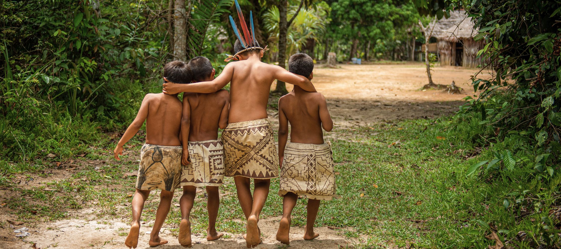 Four children walk together down a dirt path in a lush green environment, wearing traditional attire. They are barefoot and appear to be enjoying a moment of camaraderie, with one child wearing a feathered headdress. In the background, there are trees and a simple structure.