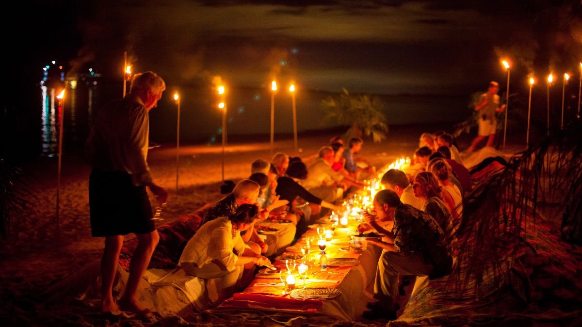 A group of people gathered around a long table on the beach at night, illuminated by candles and tiki torches. The scene features a relaxed atmosphere with guests enjoying a meal under a cloudy sky.